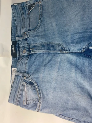 Ljusblå slitna jeans från Replay - Säljer ett par ljusblå jeans från Replay med snygga slitningar och hål på benen. Jeansen har klassisk femficksdesign, dragkedjegylf och raka ben. Perfekta för en avslappnad och trendig look.