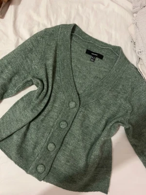 Grön kofta från H&M med stora knappar - Mysig grön kofta från H&M med v-ringning och stora matchande knappar framtill. Koftan har ribbade muddar och är tillverkad i ett mjukt, stickat material. Perfekt för lager på lager och enkel att styla till olika outfits.