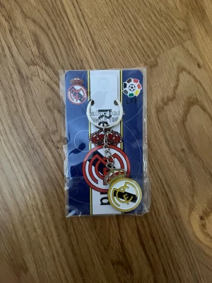 Real Madrid nyckelring - Nyckelring med Real Madrid-logga i metall och plast. Färgerna är vit, gul och svart med klubbens emblem. Perfekt accessoar för fotbollsfans som vill visa sitt stöd för laget.