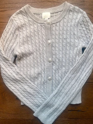 Oanvänd ljusgrå kofta - Säljer en oanvänd ljusgrå kabelstickad kofta med rund halsringning och pärlemorknappar. Koftan har långa ärmar och ribbade muddar vid ärmslut och nederkant. 🩶