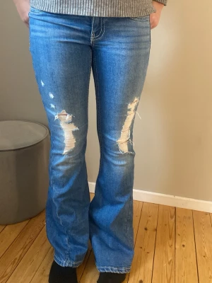 Blå bootcut jeans med slitningar - Säljer ett par blå bootcut jeans ifrån gina tricot, storlek S. Med hål på båda benen framtill. Jeansen har texturerade bakfickor med dekorativa sömmar och knappar.