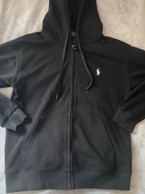 Svart hoodie från Polo Ralph Lauren - Klassisk svart hoodie med dragkedja från Polo Ralph Lauren. Har huva med dragsko, två stora fickor framtill och den ikoniska vita loggan broderad på bröstet. Tillverkad i mjukt bomullsmaterial och ribbade muddar vid ärmar och nederkant.