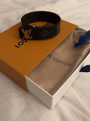 Louis Vuitton svart läderarmband - Snyggt svart armband från Louis Vuitton i präglat läder med klassiska LV-loggan i metall och monogramdetaljer. Armbandet har en stilren design och justerbart spänne. Perfekt accessoar för dig som gillar exklusiva märken och tidlös stil.