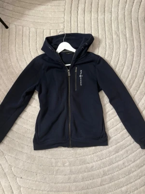 Mörkblå hoodie från Sail Racing - Mörkblå hoodie från Sail Racing med dragkedja framtill och en bröstficka med logga. Storlek 160 men motsvarar S. Tröjan har en justerbar huva med tryckt detalj och är tillverkad i ett mjukt, stretchigt material som känns skönt mot huden. Perfekt för en avslappnad och sportig stil.