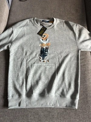 Grå sweatshirt Polo Bear Ralph Lauren - Snygg grå sweatshirt från Ralph Lauren med ikoniska Polo Bear-trycket framtill. Tröjan har rund halsringning, ribbade muddar och är tillverkad i mjukt bomullsmaterial. Perfekt för en avslappnad och trendig stil. Denna tröja är helt ny och aldrig använd. Denna tröja fungerar till storlek L samt M