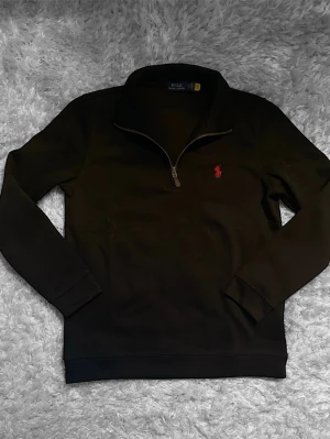 Svart halvzip-tröja från Polo Ralph Lauren - Svart långärmad tröja från Polo Ralph Lauren med halv dragkedja och klassisk krage. Tröjan har en röd broderad logga på bröstet och är tillverkad i mjukt material. Perfekt för en stilren och avslappnad look.