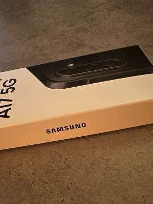 Samsung Galaxy A17 5G svart - Samsung Galaxy A17 5G är en modern Android-telefon med stilren svart design och rundade kanter. Telefonen har flera kameror på baksidan och levereras i en vit kartong med tydlig Samsung-logga. Perfekt för dig som vill ha snabb 5G och snygg look.