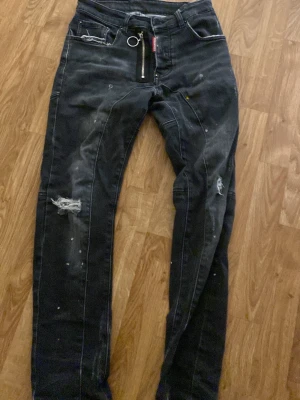 Dsquared jeans - Säljer ett par svarta jeans med slitningar och hål på benen, samt vita färgstänk för en edgy look. Jeansen har en unik dragkedjedetalj framtill och dekorativ ring. Smal passform och cool streetstil.
