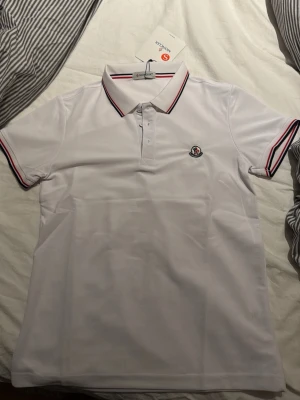 Vit pikétröja från Moncler - Vit pikétröja från Moncler med klassisk krage och knäppning framtill. Tröjan har röda och marinblå ränder på krage och ärmslut samt Moncler-logga på bröstet. Perfekt för en stilren och sportig look.