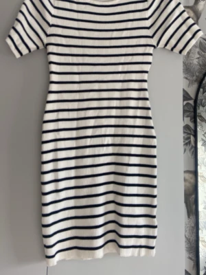Randig ribbad kortklänning - Vit och marinblå ribbad kortklänning med horisontella ränder. Klänningen har korta ärmar och en kroppsnära passform. Perfekt för en avslappnad och stilren look. Materialet känns mjukt och stretchigt, troligen bomull eller en bomullsblandning.