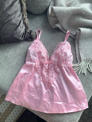 Rosa satinlinne med spetsdetaljer hjärtan - Supersött rosa linne i satin med tunna axelband och v-ringning. Framtill finns broderade spetsdetaljer i form av hjärtan och en liten rosett. Materialet är glansigt och mjukt, perfekt för en romantisk look.