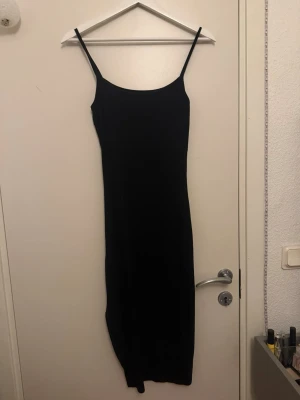 Svart ribbad långklänning - Säljer en svart långklänning med smala axelband, en liten slitt vid benet och ribbad struktur. Klänningen har en kroppsnära passform och är tillverkad i ett mjukt, stretchigt material som känns bekvämt mot huden. Perfekt för dig som gillar en enkel men stilren look.