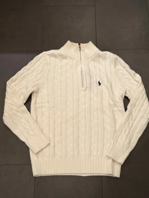 Vit kabelstickad tröja Polo Ralph Lauren - Kabelstickad vit tröja från Polo Ralph Lauren med halv dragkedja och hög krage. Tröjan har långa ärmar, ribbade muddar och den klassiska broderade loggan på bröstet. Perfekt för dig som gillar stilren och tidlös design.