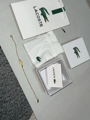 Guldfärgat armband från Lacoste - Stilrent guldfärgat armband från Lacoste med den ikoniska krokodil-loggan som hänge. Tunt kedjearmband i metall med minimalistisk design. Levereras med originalask, påse och kort från Lacoste.