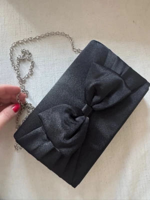 Svart kuvertväska med rosett - Snygg svart kuvertväska i satinliknande material med dekorativ rosett på framsidan. Väskan har en silverfärgad kedja som axelrem och stängs med magnetknapp. Perfekt för att lyfta din outfit med en elegant touch. Har bundlerabatt vid köp av fler varor🥰