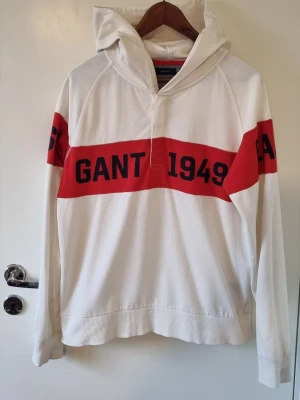 Vit hoodie från Gant med röd detalj - Vit hoodie från Gant med bred röd rand över bröstet och armarna där det står 'GANT 1949' och 'CONNECTICUT' i svart text. Hoodien har  knappar vid halsen och en stor huva. Tillverkad i mjukt bomullsmaterial, perfekt för en avslappnad och sportig stil. Storlek 176