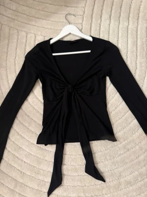 Svart långärmad knytblus - Trendig svart långärmad topp med djup v-ringning och knyt framtill. Sitter snyggt och har en croppad passform som passar perfekt till höga jeans eller kjol. Enkel men ändå stylish design som funkar till många olika looks.