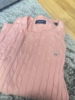 Rosa kabelstickad tröja från GANT - Snygg rosa kabelstickad tröja från GANT med rund halsringning och diskret broderad logga på bröstet. Tröjan har klassisk kabelstickad struktur och ribbade muddar vid ärmslut och nederkant. Perfekt för dig som gillar preppy stil.