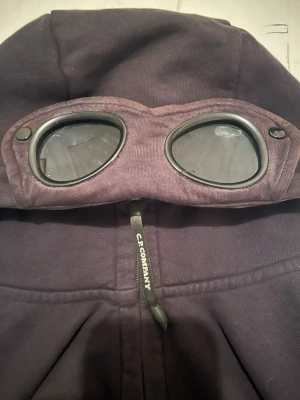 Svart hoodie med goggles från C.P. Company - Säljer en svart hoodie från C.P. Company med ikoniska inbyggda goggles i huvan och dragkedja framtill. Tillverkad i mjukt bomullstyg och har en sportig streetwear-look. Perfekt för dig som vill sticka ut med en unik detalj på din hoodie.