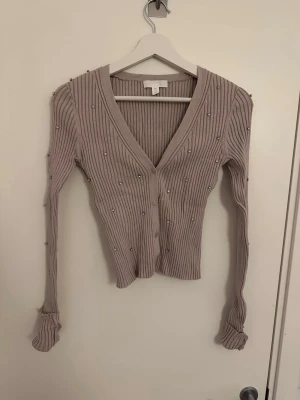 Beige ribbad kofta med pärlor - Säljer en beige ribbad kofta med v-ringning och dekorativa pärlor över hela plagget. Koftan har långa ärmar, smal passform och knäppning framtill. Perfekt för dig som gillar detaljer och en stilren look.
