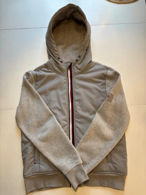 Grå hoodiejacka från Tommy Hilfiger - Grå hoodiejacka från Tommy Hilfiger med dragkedja framtill och klassiska Hilfiger-detaljer på ärm och rygg. Jackan har långärmade ärmar och huva. Perfekt för lager-på-lager och chill stil. Storlek M. Skick 9/10.