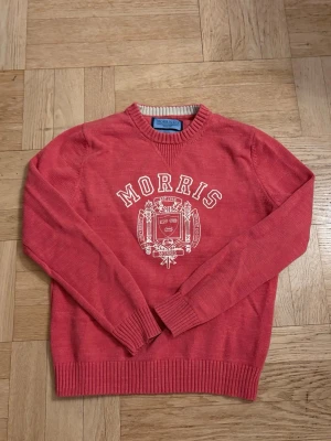 Röd stickad tröja från Morris - Röd stickad tröja från Morris med rund halsringning och ribbade muddar. Framtill finns ett stort vitt tryck med Morris-logga och emblem. Tröjan har klassisk passform och är perfekt för dig som gillar preppy stil.