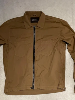 Brun overshirt från Oskar Jacobsson - Stilren brun overshirt från Oscar Jacobsson med två bröstfickor och svart dragkedja framtill. Den är endast använd vid 2 tillfällen. Den har inga defekter. Bara höra av sig vid mista fundering🤗