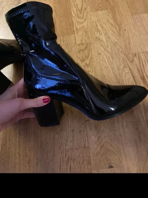 Svarta lackade stövletter med klack - Säljer ett par glansiga svarta stövletter i lackmaterial med spetsig tå och bred klack. Skorna har en tight passform runt ankeln.