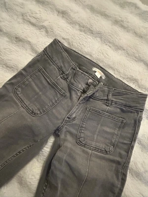 Grå jeans med stora fickor - Snygga grå boot cut jeans med stora fyrkantiga fickor fram. Jeansen har dubbla knappar i midjan och klassiska bakfickor. Perfekta för en avslappnad och trendig look. Materialet är mjukt denim som ger en skön känsla hela dagen. 