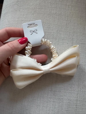 Krämvit scrunchie med rosett - Snygg scrunchie i satinliknande material med stor krämvit rosett. Perfekt för att piffa till håret med en elegant touch. Elastisk och mjuk, passar till många frisyrer. Enkel att matcha med olika outfits. Har bundlerabatt vid köp av fler varor🥰