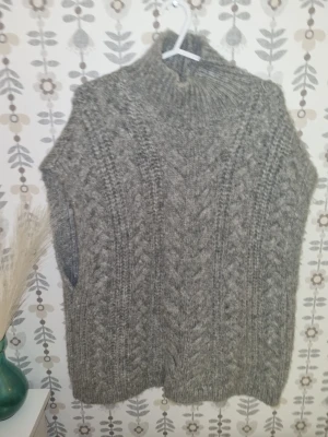 Grå stickad poncho med polokrage - Säljer en grå stickad poncho med polokrage och grovt kabelstickat mönster. Ponchon är ärmlös och har en avslappnad, oversized passform. Perfekt att slänga över en tröja för extra värme och stil.