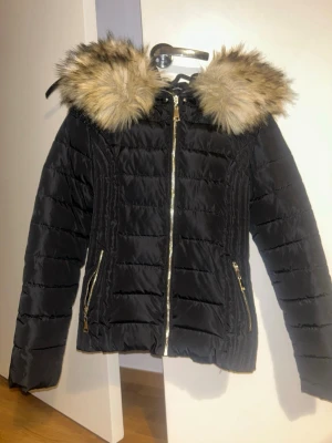 Svart dunjacka med päls - Snygg svart dunjacka med stor luva kantad av beige fuskpäls. Jackan har guldfärgade dragkedjor och två fickor framtill. Quiltad design som ger en schysst passform och extra värme. Perfekt för kalla vinterdagar och riktigt trendig look. Helt ny aldrig använt fick den som present men inte min still. Pris kan diskuteras 