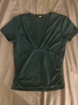 Mörkgrön topp med v-ringning BikBok - Snygg mörkgrön topp från BikBok med djup v-ringning och korta ärmar. Figurnära passform med rynkade detaljer i sidorna som ger en cool siluett. Perfekt att styla med jeans eller kjol för en trendig look.
