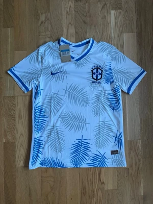 Vit Brasilien fotbollströja Nike - Snygg vit Brasilien fotbollströja från Nike med blå palmbladsmönster och blå detaljer vid krage och ärmslut. Tröjan har officiellt Brasilien-emblem på bröstet och är tillverkad i lätt, ventilerande material som passar perfekt till träning eller match.