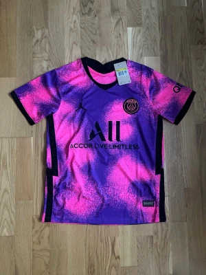 PSG Mbappé matchtröja rosa/lila - Säljer en PSG matchtröja i samarbete med Jordan, med Mbappé och nummer 7 på ryggen. Tröjan har en unik rosa och lila färgkombination med svarta detaljer, klubbmärke och sponsortryck framtill. Tillverkad i lätt och ventilerande funktionsmaterial, perfekt för fotboll.
