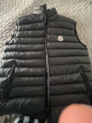 Svart dunväst från Moncler - Snygg svart dunväst från Moncler med hög krage och dragkedja framtill. Västen har två sidofickor med dragkedja och Moncler-logga på bröstet. Perfekt för lager-på-lager och håller dig varm under kyliga dagar.