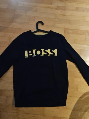 Svart sweatshirt från BOSS - Svart sweatshirt från BOSS med stort gult logotryck på bröstet. Klassisk rund halsringning och långa ärmar. Perfekt för en stilren och avslappnad look.