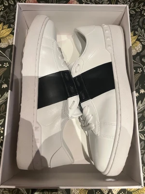 Valentino Garavani open vit blåa  - Snygga vita sneakers från Valentino med ett brett blåt band över mitten. Skorna har låga kanter, klassisk siluett och detaljerade sömmar. Tillverkade i slätt läder med vit sula och diskreta nitar på sidan för en cool touch.