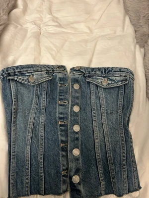 Blå jeanstopp med knappar från Zara - Trendig blå jeanstopp från Zara med raka sömmar, dekorativa fickor framtill och silverfärgade knappar längs hela framsidan. 