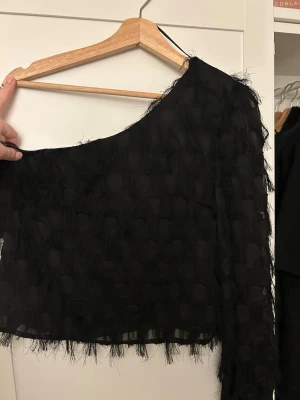 Svart oneshoulder topp Gina Tricot - Säljer en svart oneshoulder topp från Gina Tricot med fransiga detaljer och lätt transparent tyg. Toppen har en unik struktur med diskreta prickar💞