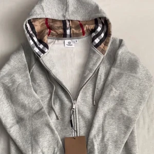 Burberry zipper/kofta - Snygg grå hoodie från Burberry med dragkedja framtill och klassiskt rutmönster i huvan. Tillverkad i mjukt bomullsmaterial och har justerbara snören vid huvan. Perfekt för en avslappnad och trendig look.