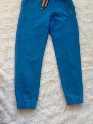 Blå mjukisbyxor med dragsko - Blå mjukisbyxor med orange dragsko i midjan och resår vid bensluten. Liten broderad detalj på ena benet. Sköna och avslappnade, perfekta för chill dagar. Tillverkade i mjukt bomullstyg.