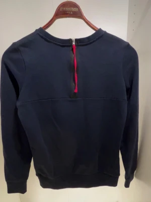 Mörkblå Sail Racing sweatshirt - Mörkblå sweatshirt från Sail Racing med stor vit text på bröstet. Tröjan har rund halsringning, långa ärmar och en cool dragkedja med röd detalj i nacken. Perfekt för dig som gillar sportig och avslappnad stil.