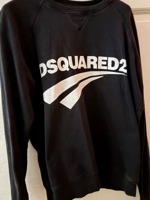  Dsquared2 sweatshirt - Tidlös svart sweatshirt från Dsquared2 med stor vit logga och grafiskt tryck på bröstet. Plagget är från deras vintage-edition, vilket ger plagget ett naturligt ”slitage”, utan att pruta på kvaliteten .Tröjan har rund halsringning och långa ärmar. Inköpt januari 2025 för 2499:-.