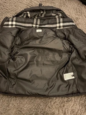 Svart dunjacka från Burberry - Snygg svart dunjacka från Burberry med klassiskt rutigt foder i grått, vitt och svart. Jackan har en vadderad insida och detaljer som ger en exklusiv känsla. Perfekt för kalla vinterdagar och har en tidlös design.