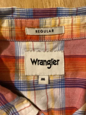 Rutig kortärmad skjorta i bomull - Kortärmad skjorta med rutigt mönster i orange, blått, gult och vitt. Skjortan har klassisk krage, knappar framtill och en liten broderad detalj på bröstet. Tillverkad i tunn bomull, perfekt för varma dagar.