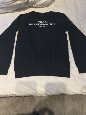 Svart sweatshirt Peak Performance - Snygg svart sweatshirt från Peak Performance med vit logga och text på bröstet. Klassisk rund halsringning och ribbade muddar vid ärmar och nederkant. Perfekt för en avslappnad och stilren look.