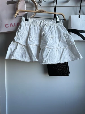 Vit volangskort med resårmidja - Supersöt vit skort med volanger och råa kanter. Skorten har elastisk midja och är gjord i ett lätt, mjukt tyg som ger en flowy känsla. Perfekt för dig som vill ha kjol-looken men shortsens bekvämlighet. Passar till många olika stilar!