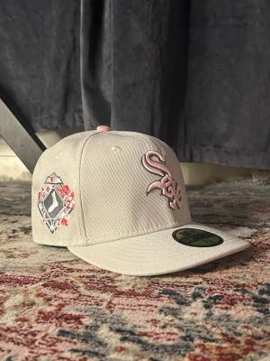 Vit New Era White Sox keps - Snygg vit keps från New Era med White Sox-logga broderad i ljusrosa framtill. Kepsen har rak skärm och detaljer i rosa, samt en unik broderad patch med blommor på sidan. Tillverkad i slitstark bomull. Perfekt för dig som vill sticka ut med stilren streetwear.