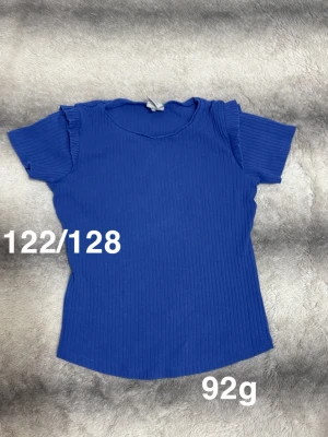 Blå ribbad topp med volangärm - Blå ribbad topp med korta ärmar och volangdetaljer vid axlarna. Rund halsringning och stretchigt material som ger en bekväm passform. Perfekt för dig som gillar en enkel men ändå söt stil.🌸 Fint använt skick. Storlek 122/128 